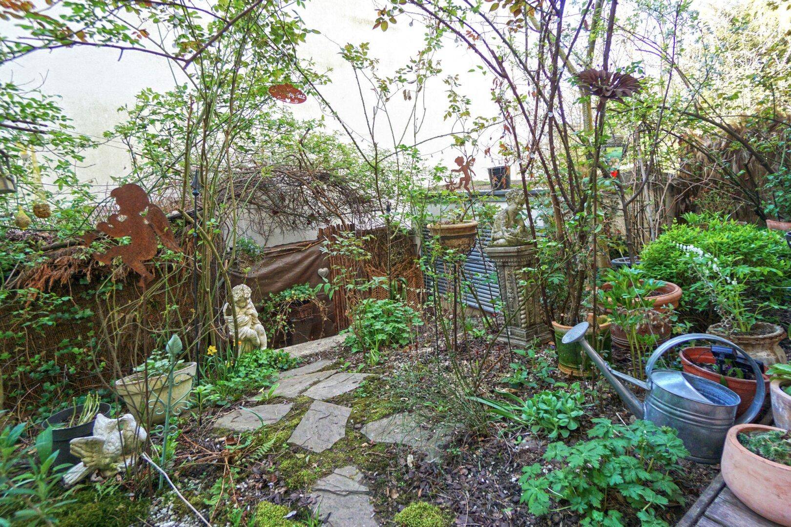 Verwunschener Gartenbereich mit Steinpfad, üppiger Vegetation und dekorativen Elementen.