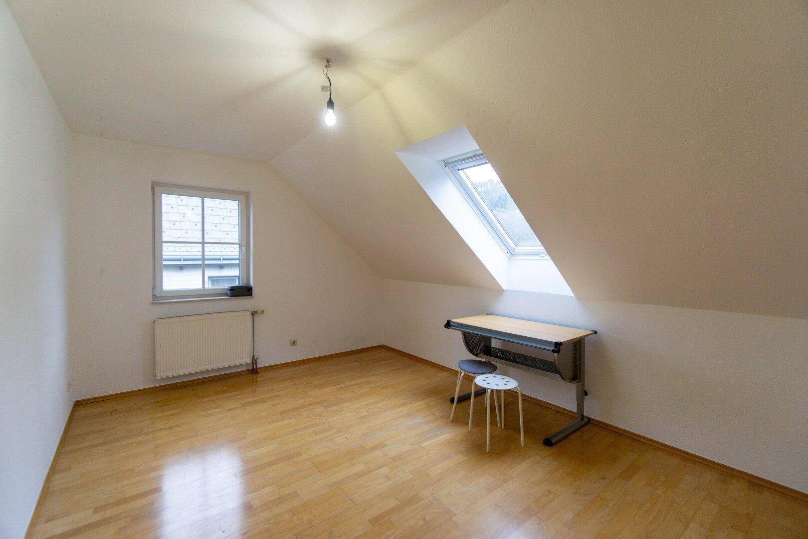 Helles Zimmer im Dachgeschoss mit Parkettboden, Dachfenster und Schreibtisch.
