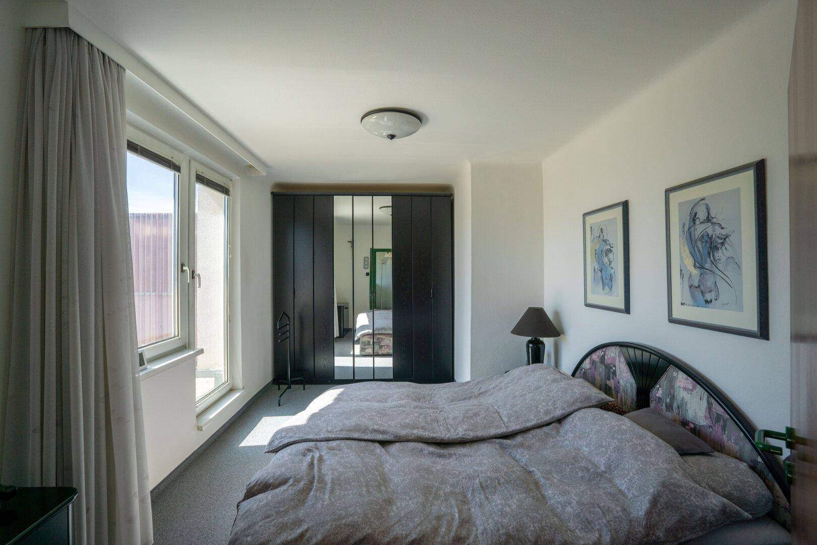 Schlafzimmer mit Doppelbett, großem Spiegelschrank und hellem Tageslicht durch die Fenster.