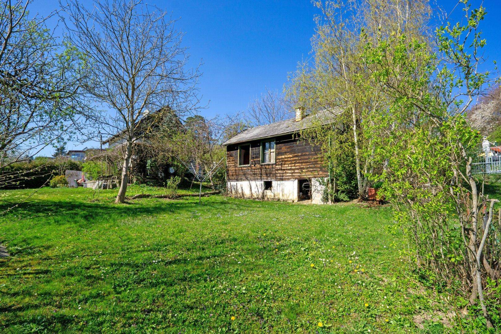 Großer Gartenbereich mit Rasenfläche und einem rustikalen Holzhaus im Hintergrund unter blauem Himmel.