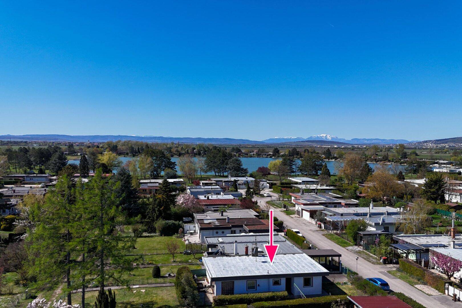 Luftaufnahme des Hauses mit Flachdach, umgeben von Gärten und dem See im Hintergrund.