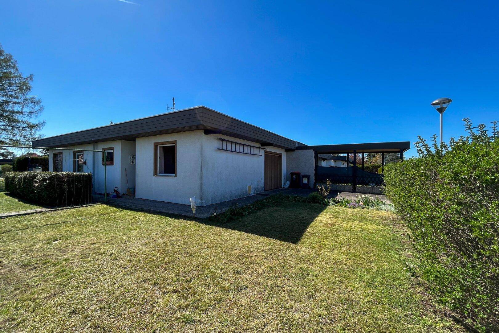 Ansicht des Hauses von der Seite mit Rasenfläche, Hecke und Carport unter blauem Himmel.