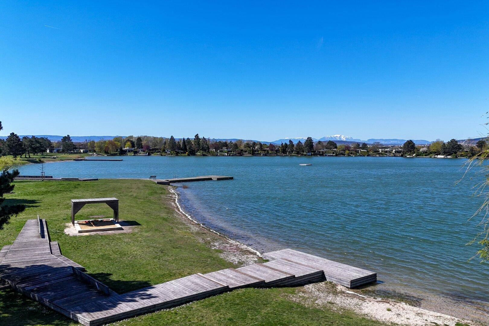 Weitläufiger Seeuferbereich mit Holzstegen, Liegewiese und klarem Wasser.