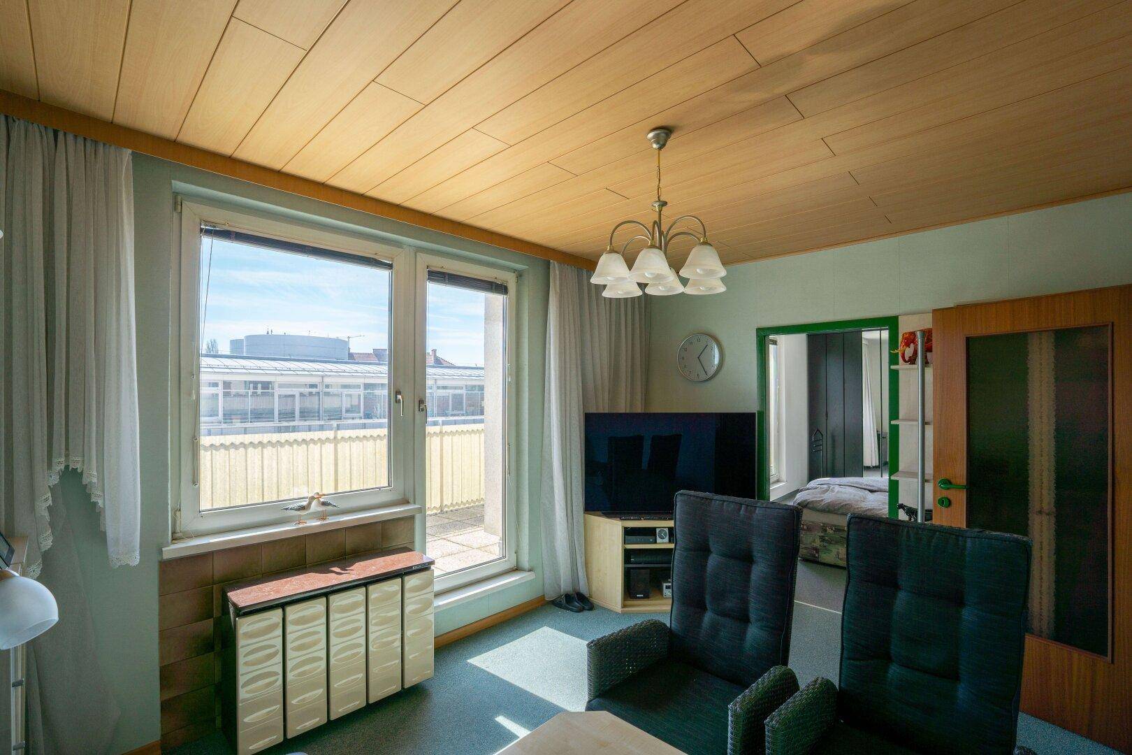 Helles Wohnzimmer mit Holzdecke, Teppichboden und Zugang zur Terrasse durch große Fenster.
