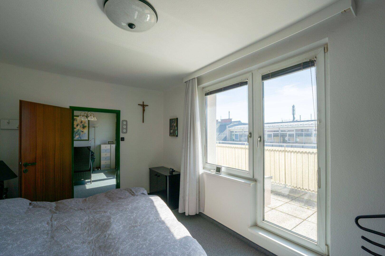 Schlafzimmer mit Zugang zur Terrasse, hellem Tageslicht und Blick auf die umliegenden Gebäude.