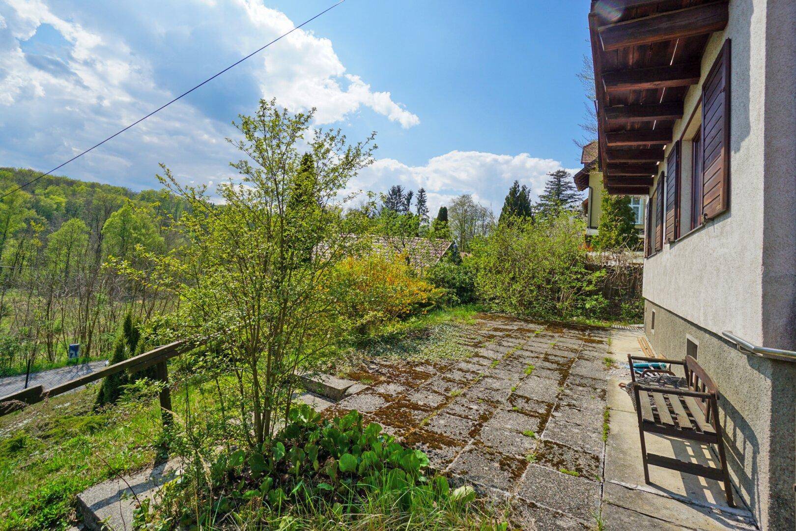 Sonniger Terrassenbereich neben dem Haus mit einer Holzbank und Blick ins Grüne.