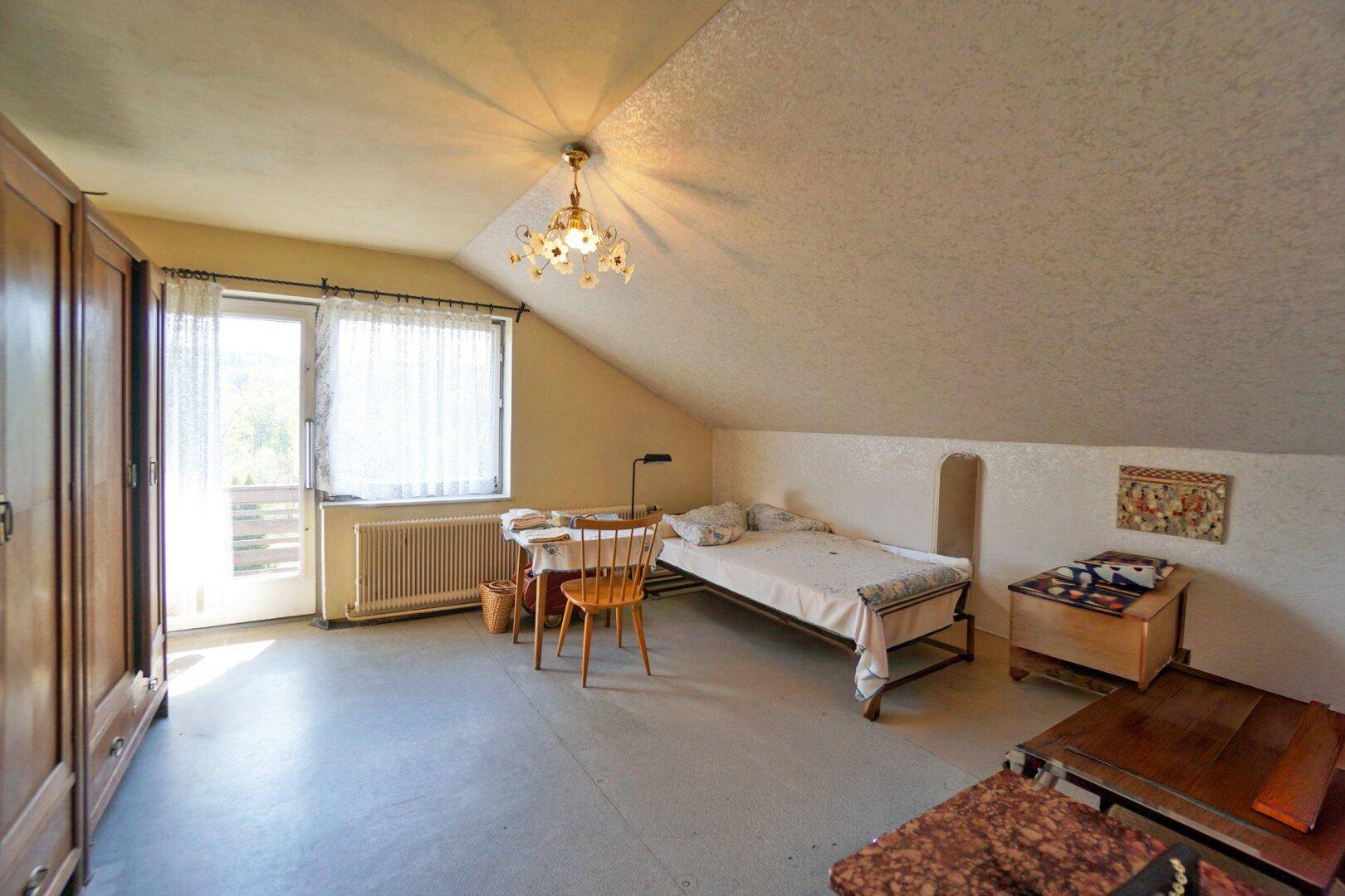 Helles Schlafzimmer im Dachgeschoss mit schrägen Wänden, Balkonzugang und einfacher Einrichtung.