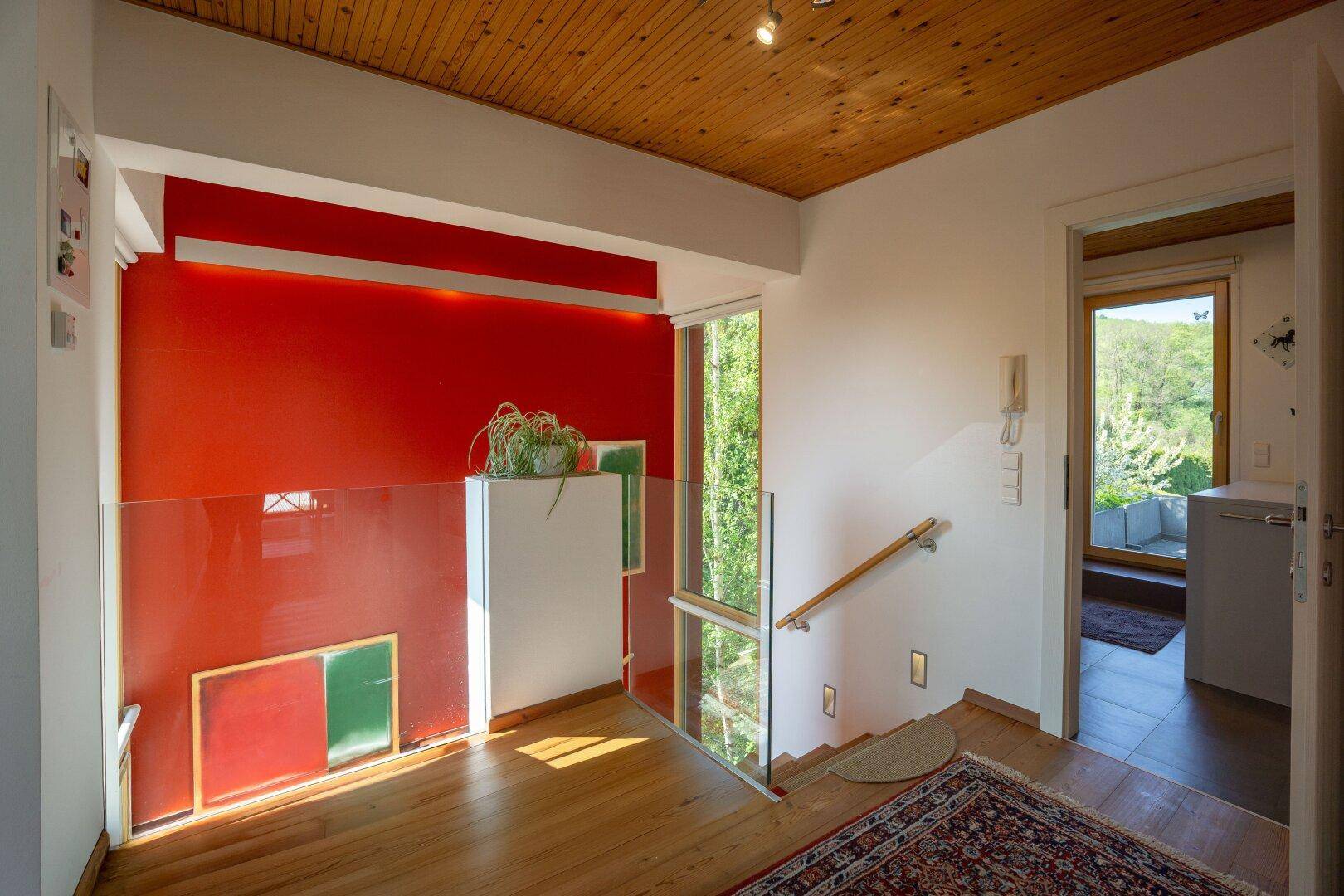 Moderner Treppenaufgang mit Glasgeländer, roter Akzentwand und Holzboden.