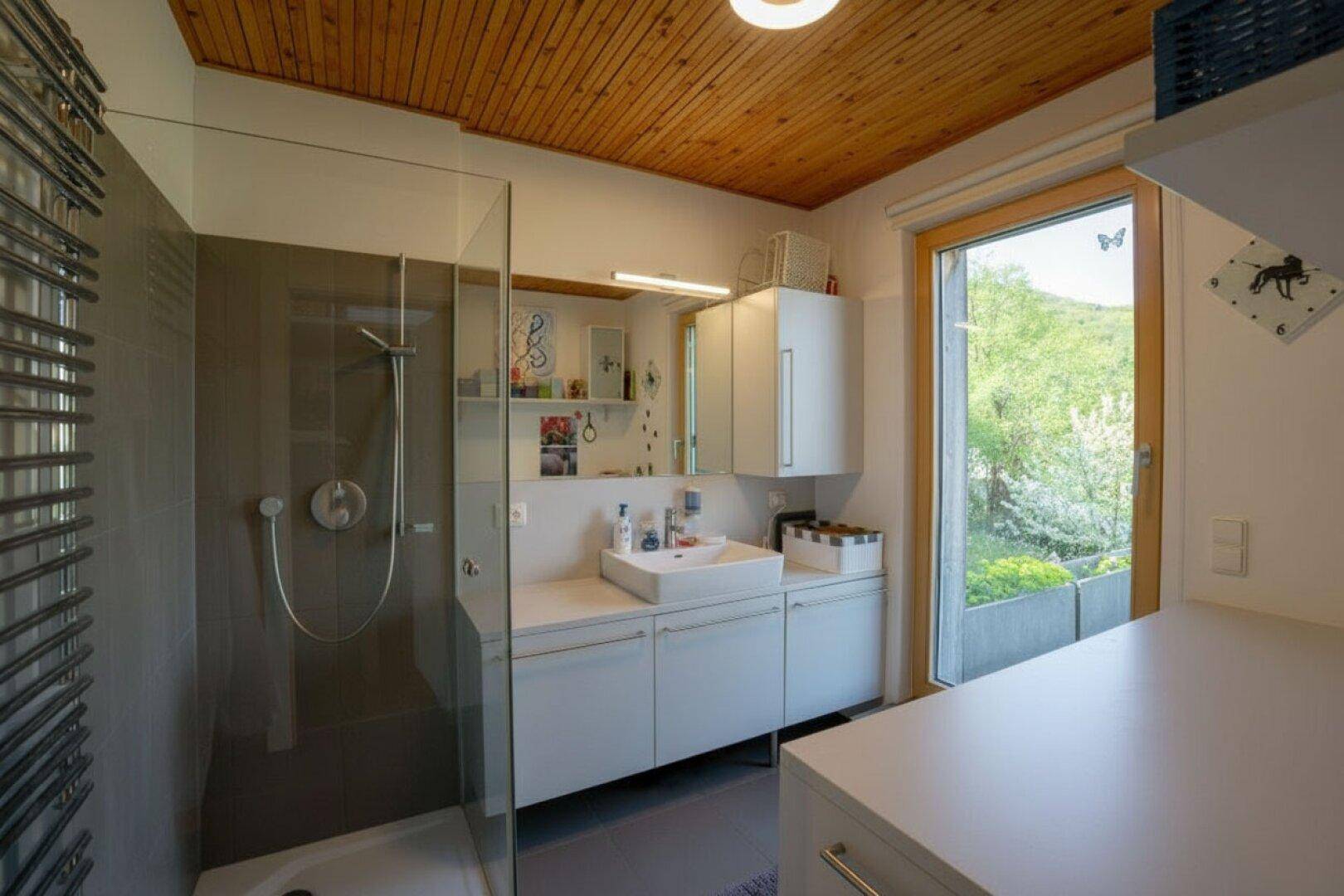 Modernes Badezimmer mit Duschkabine, Holzdecke und Fenster mit Blick ins Grüne.