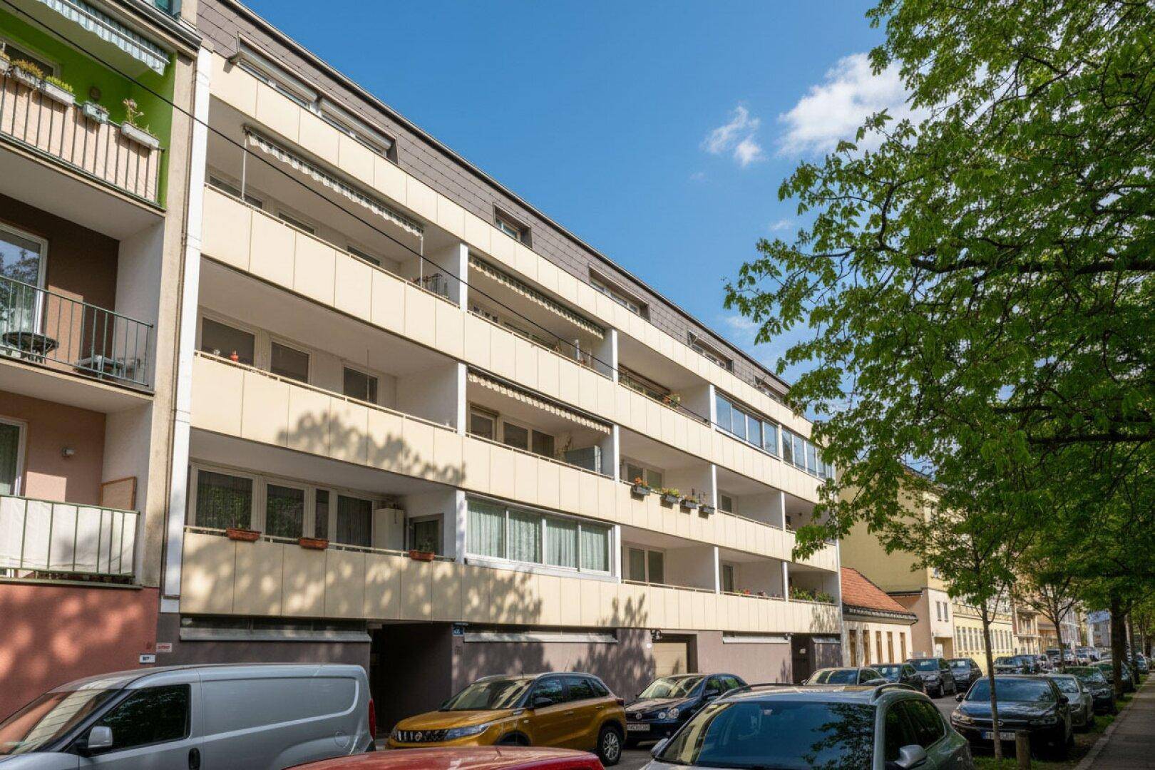 Mehrfamilienhaus mit mehreren Balkonen und parkenden Autos auf der Straße.