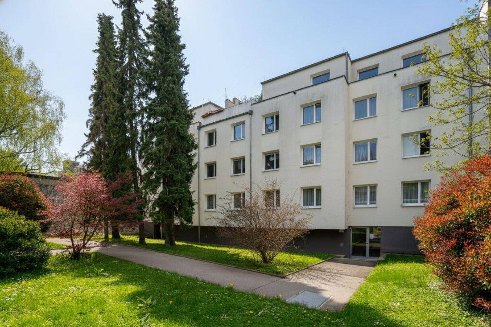 Wohngebäude mit gepflegter Grünfläche und Bäumen im Vordergrund unter blauem Himmel.