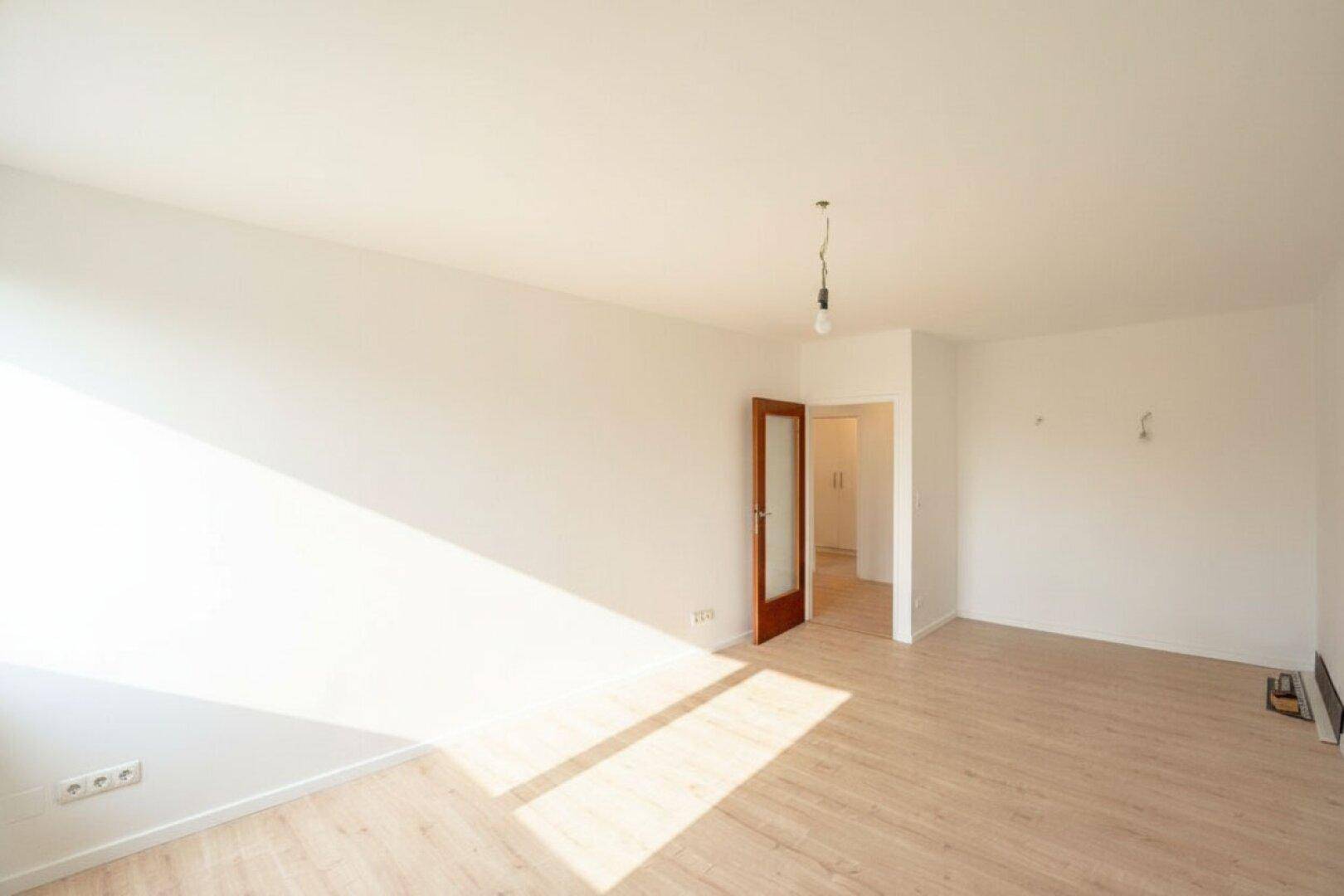 Lichtdurchfluteter Raum mit hellem Laminatboden und einer Holztür, die in einen Flur führt.