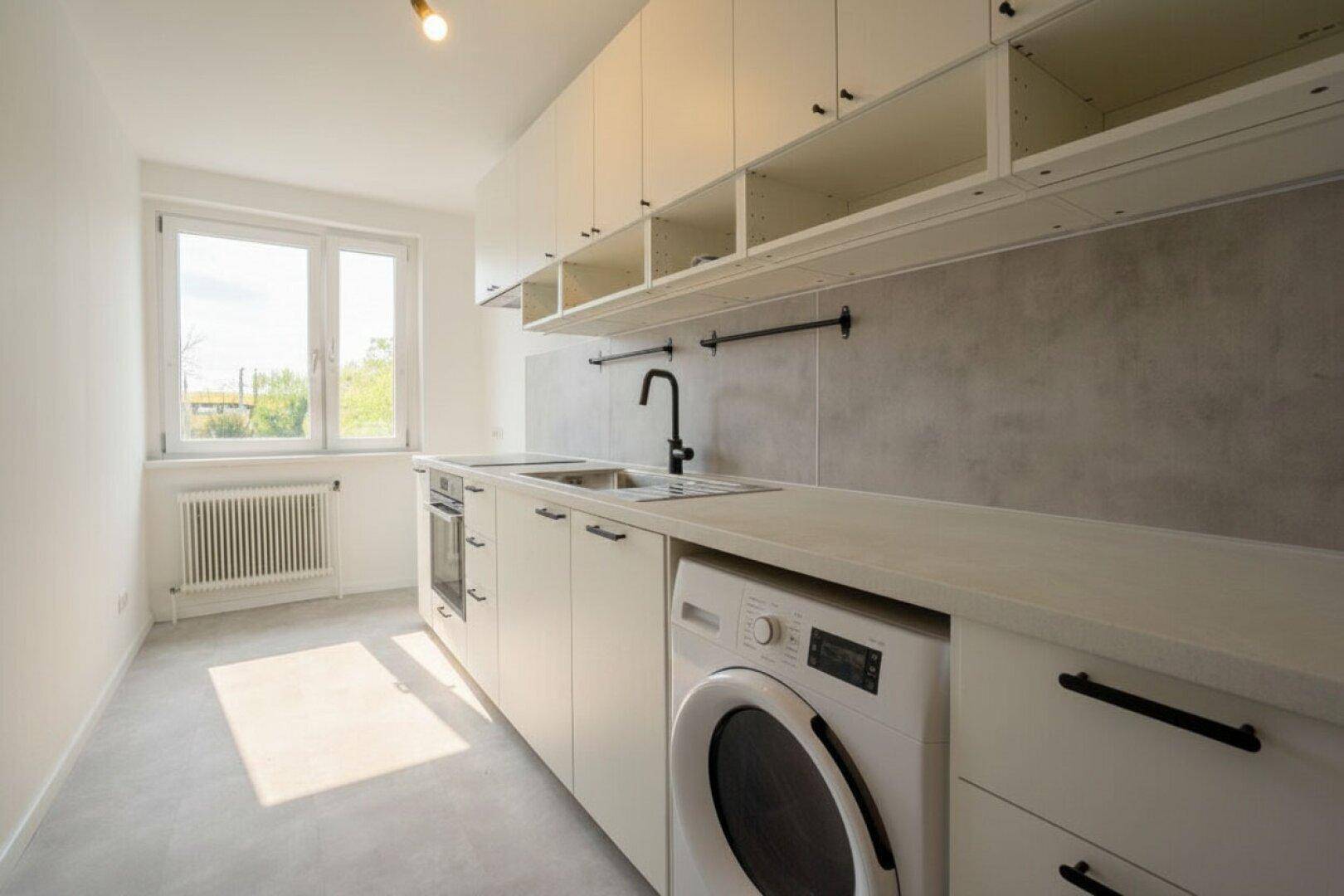 Moderne Küche mit weißen Schränken, integrierten Geräten und Waschmaschine unter dem Fenster.