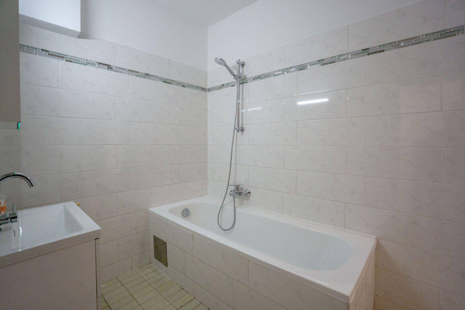 Badezimmer mit weißer Badewanne, Duscharmatur und hellen Fliesen an Wänden und Boden.