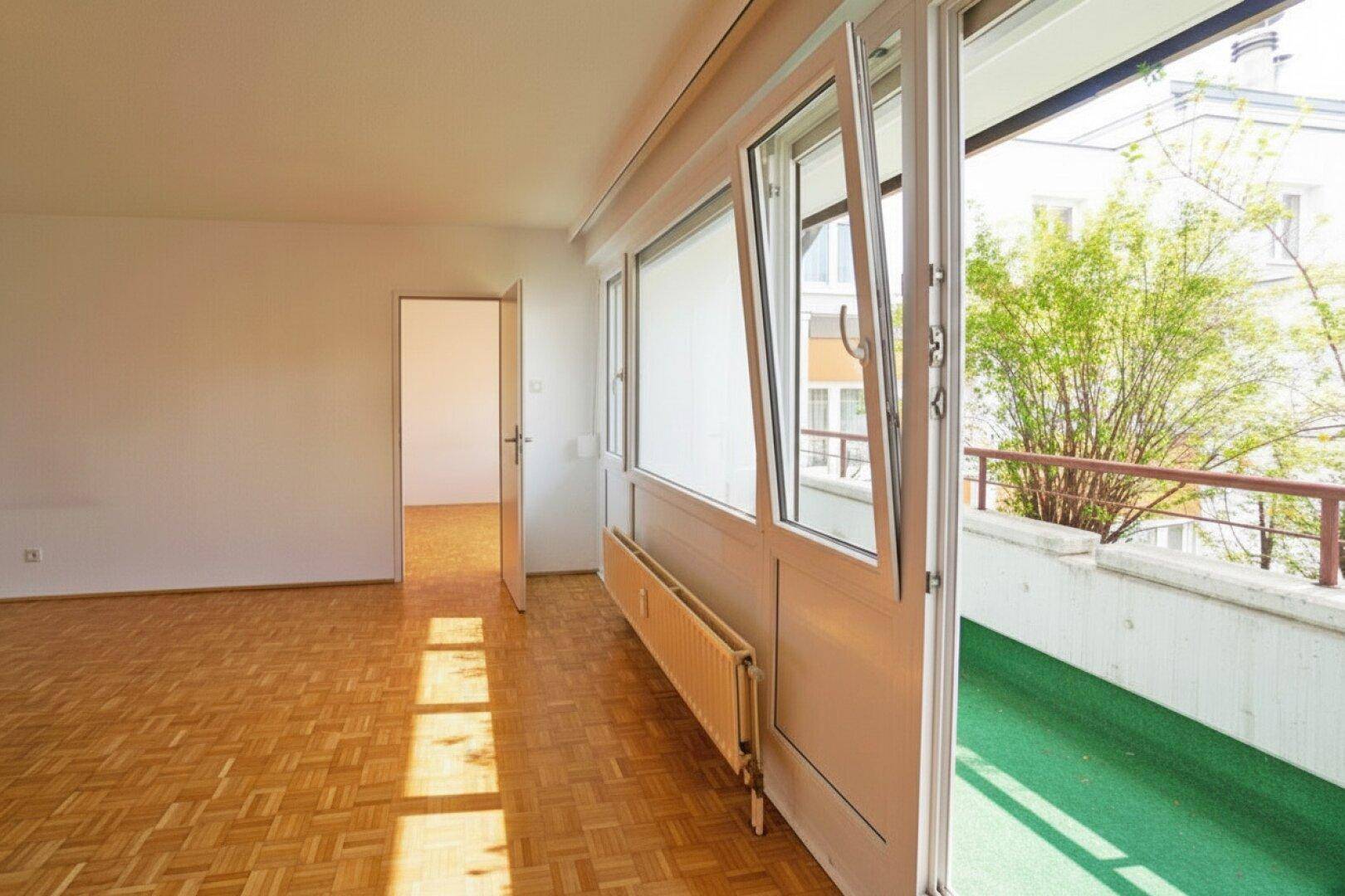 Wohnzimmer mit Parkettboden und geöffneter Balkontür, die den Blick ins Grüne freigibt.