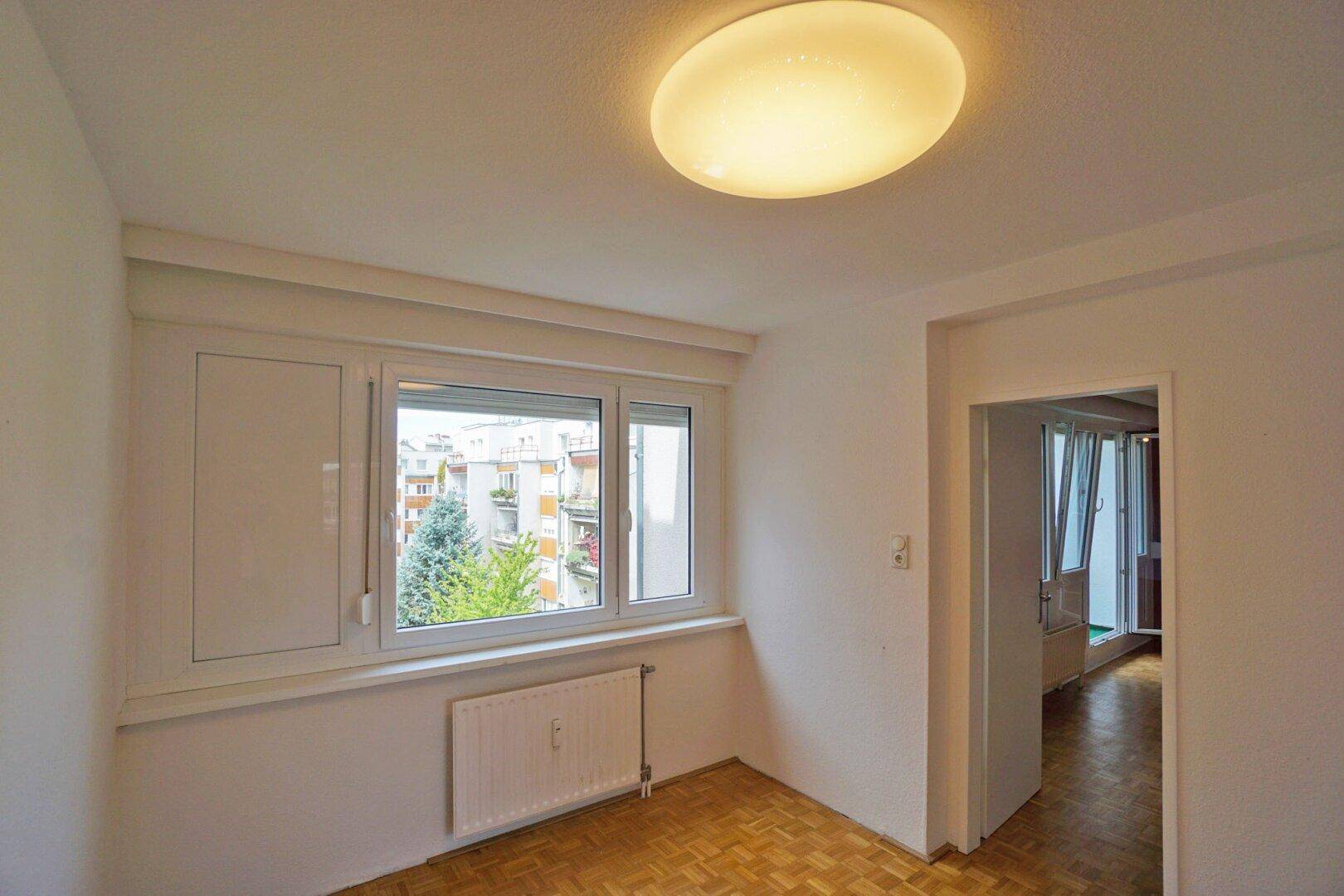 Helles Schlafzimmer mit Parkettboden und Fenster mit Blick auf den grünen Innenhof.