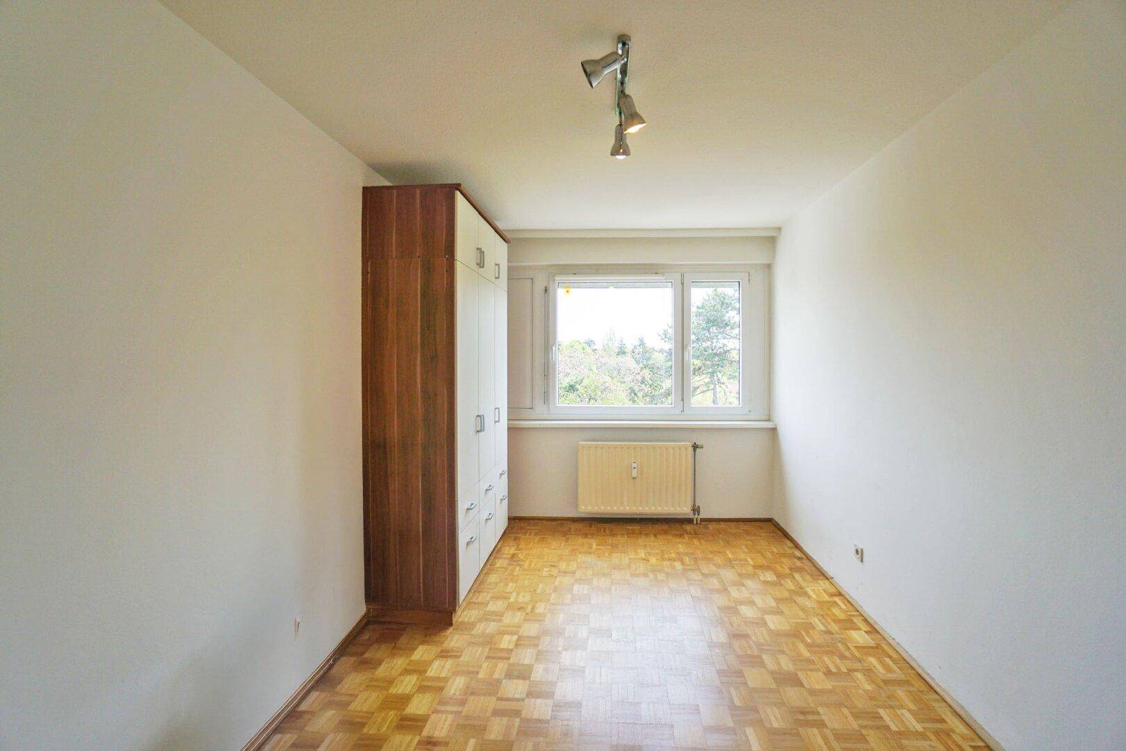 Schlafzimmer mit Parkettboden, großem Kleiderschrank und Fenster mit Blick ins Grüne.
