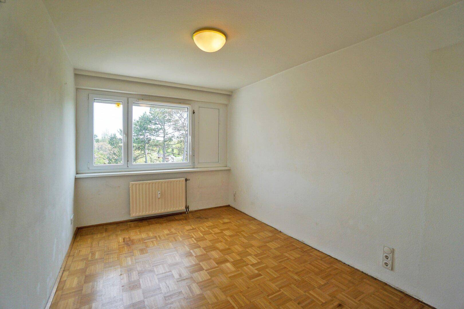 Ruhiges Schlafzimmer mit Parkettboden und Fenster mit Blick auf die grüne Umgebung.
