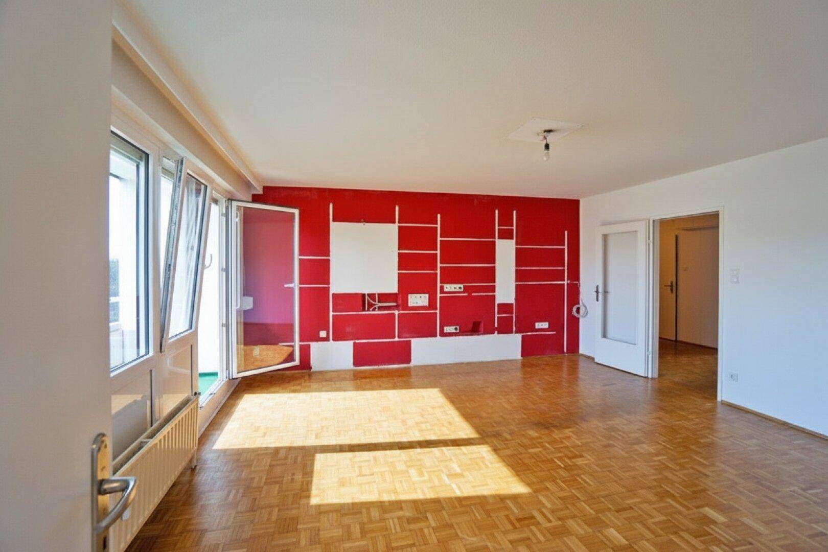 Geräumiges Wohnzimmer mit Parkettboden, roter Akzentwand und Zugang zum Balkon.