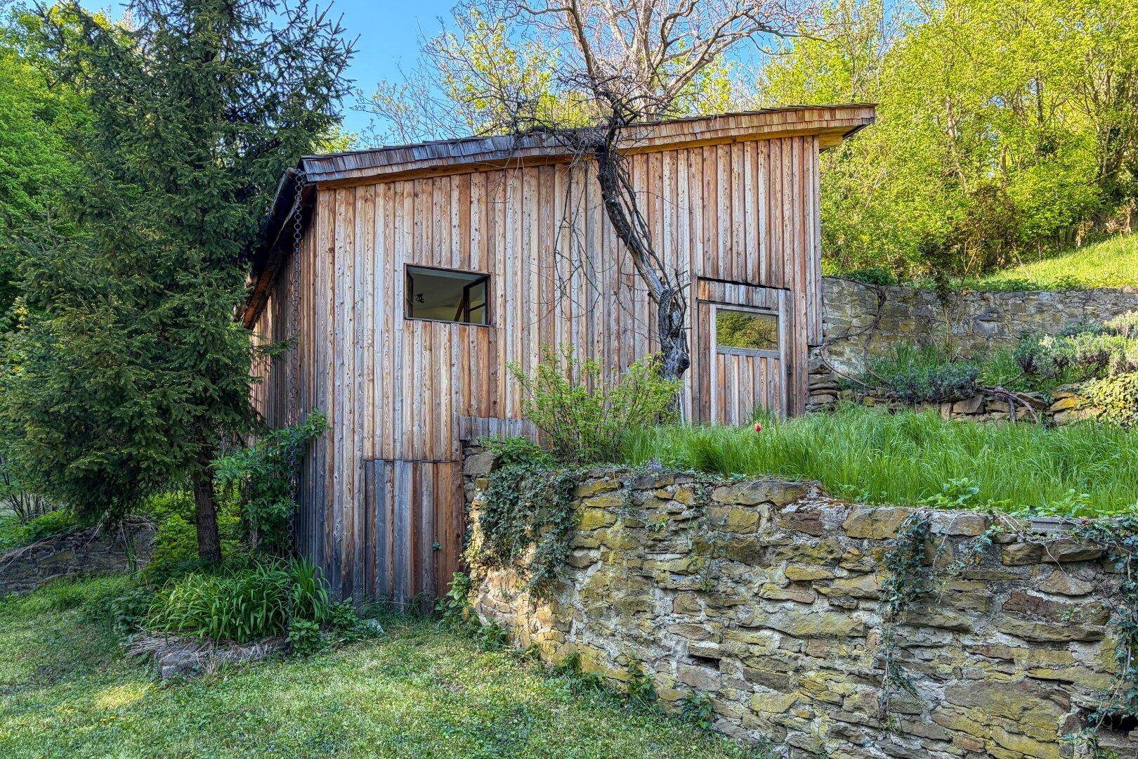 Rustikales Holzgebäude neben einer alten Steinmauer, umgeben von üppigem Grün.