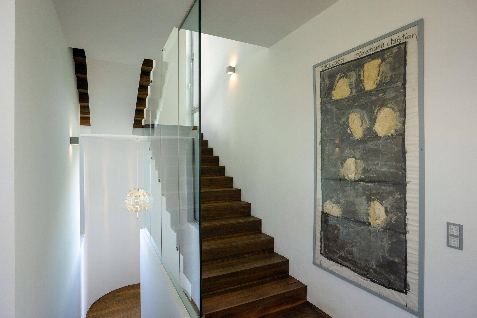 Moderne Holztreppe mit Glasgeländer und Kunstwerk an der Wand in einem hellen Treppenhaus.