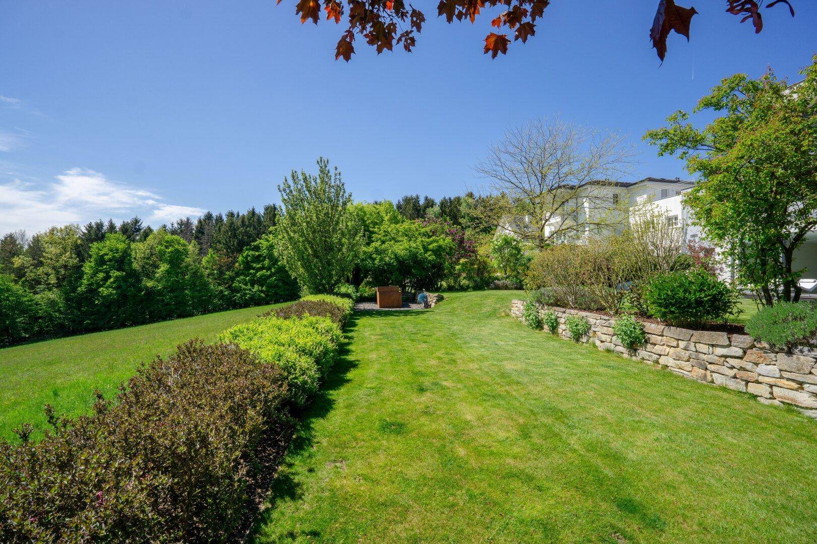 Weitläufiger, gepflegter Garten mit Rasenflächen, Sträuchern und einem Steinwall.