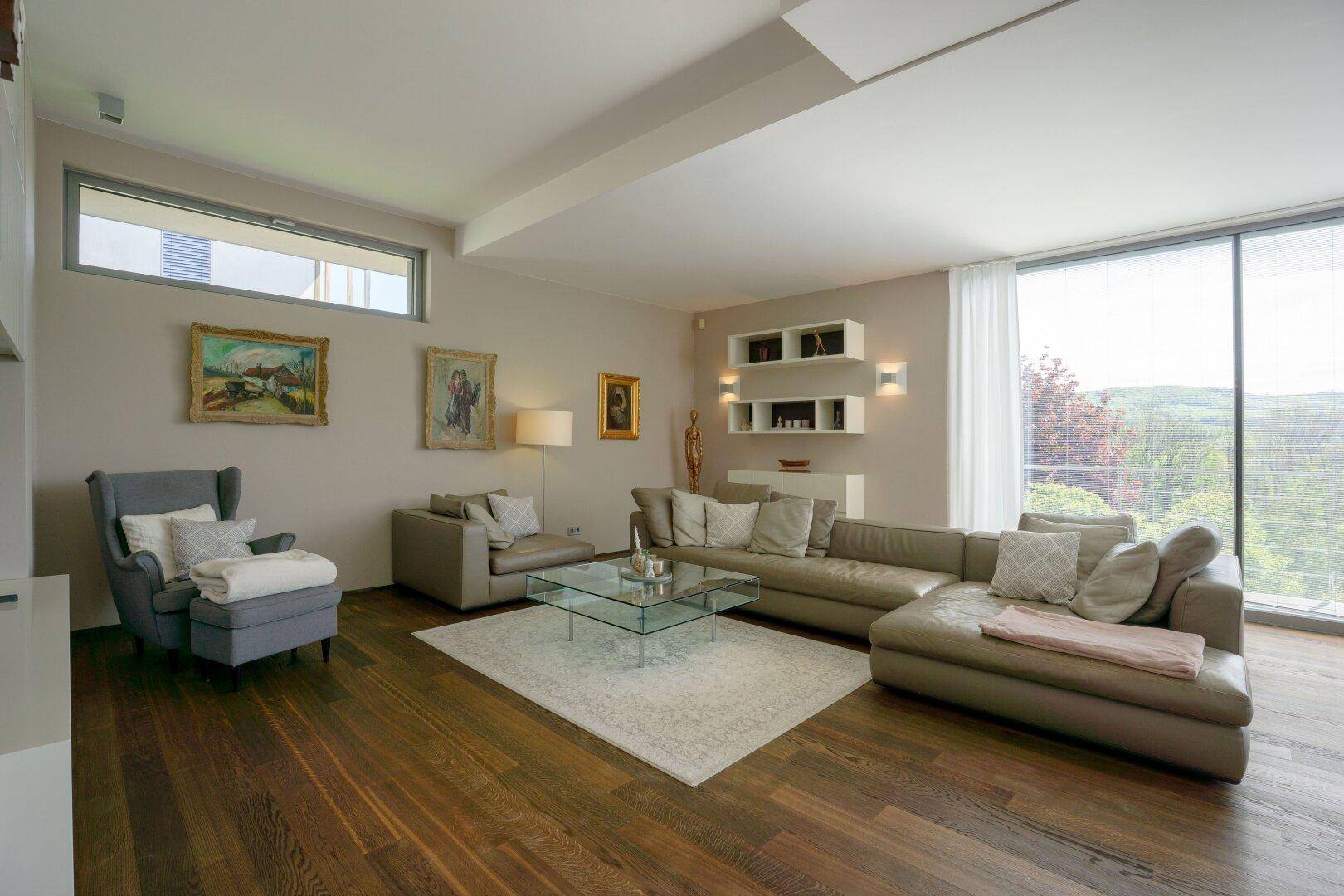 Helles Wohnzimmer mit beigem Sofa, Holzboden und großen Fenstern mit Landschaftsblick.
