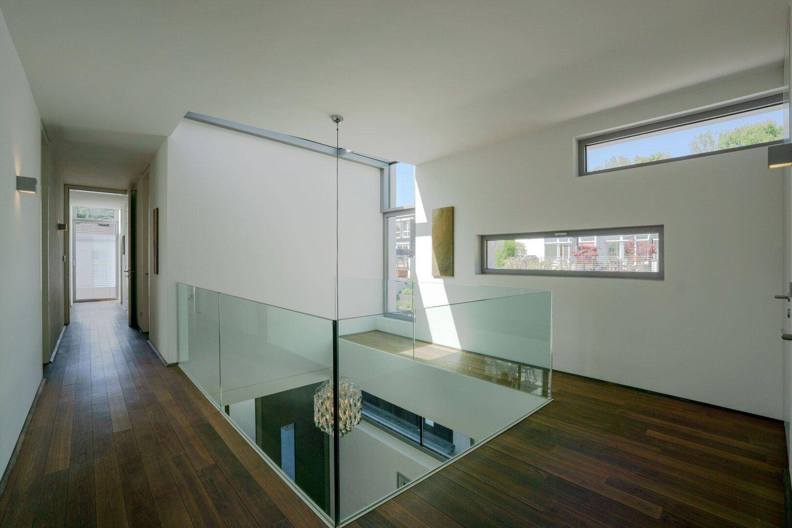 Heller Flur im Obergeschoss mit Glasgeländer, Holzboden und Fenstern mit Ausblick.