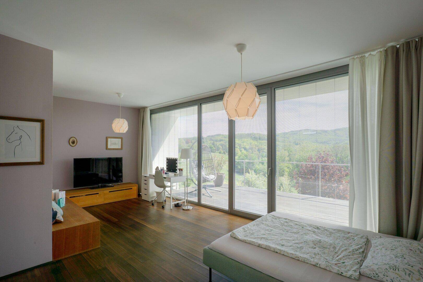 Geräumiges Schlafzimmer mit Holzboden, Schreibtisch und großen Fenstern mit Zugang zur Terrasse.