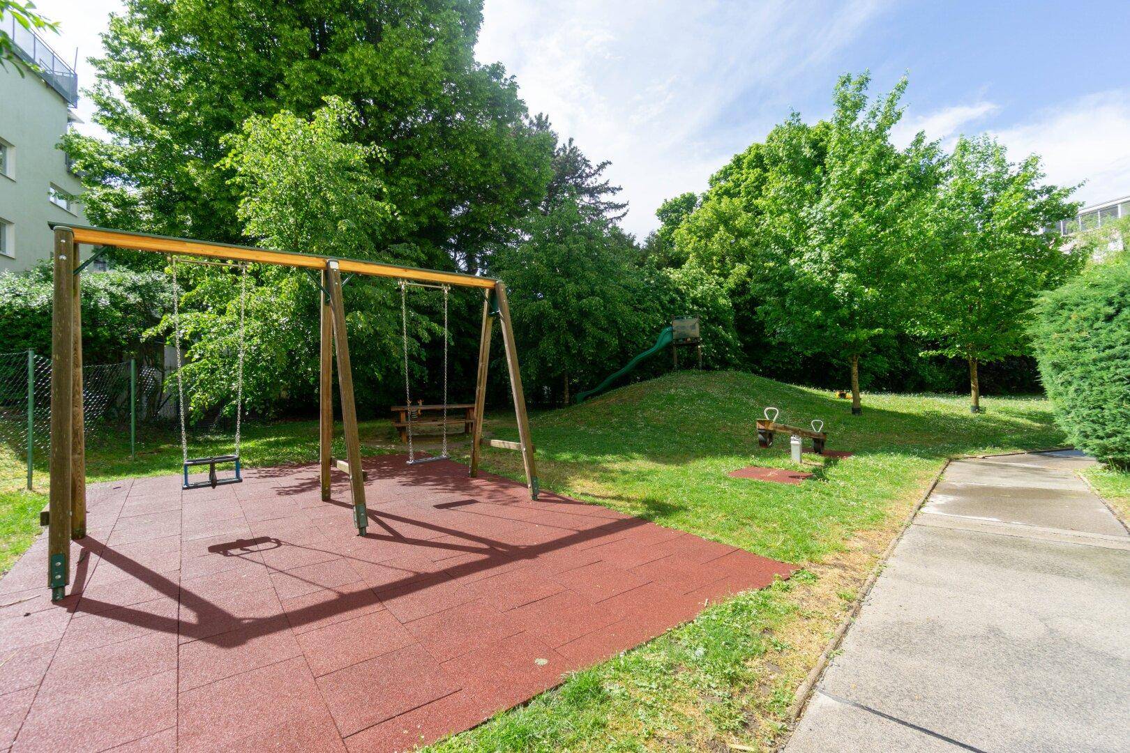 Kinderspielplatz mit Schaukeln, Rutsche und Wippe, umgeben von Bäumen und Grünflächen.