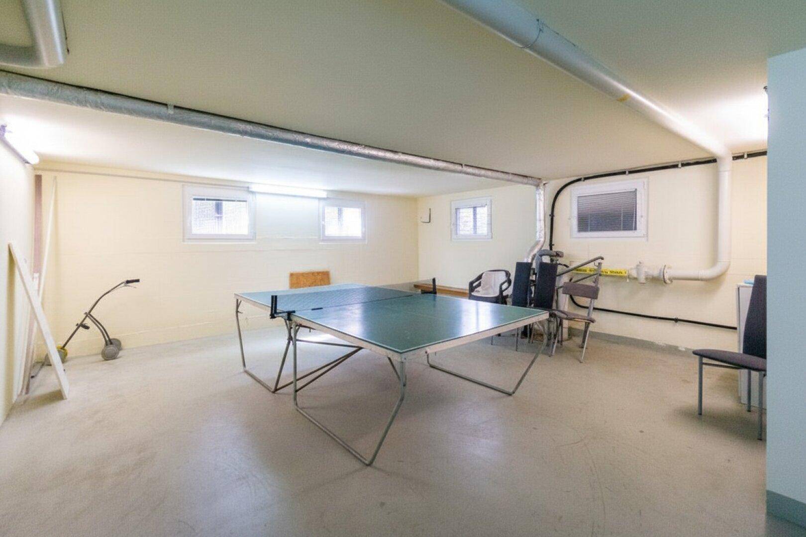 Geräumiger Kellerraum mit Tischtennisplatte, kleinen Fenstern und sichtbaren Versorgungsleitungen.