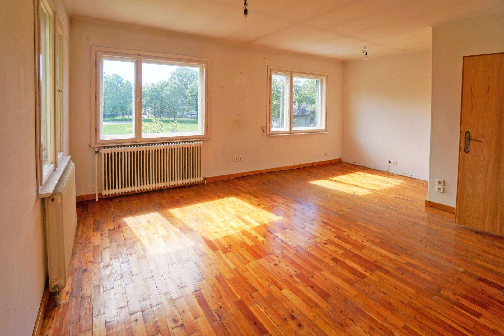 Geräumiges Zimmer mit Holzboden, zwei Fenstern und Blick ins Grüne.
