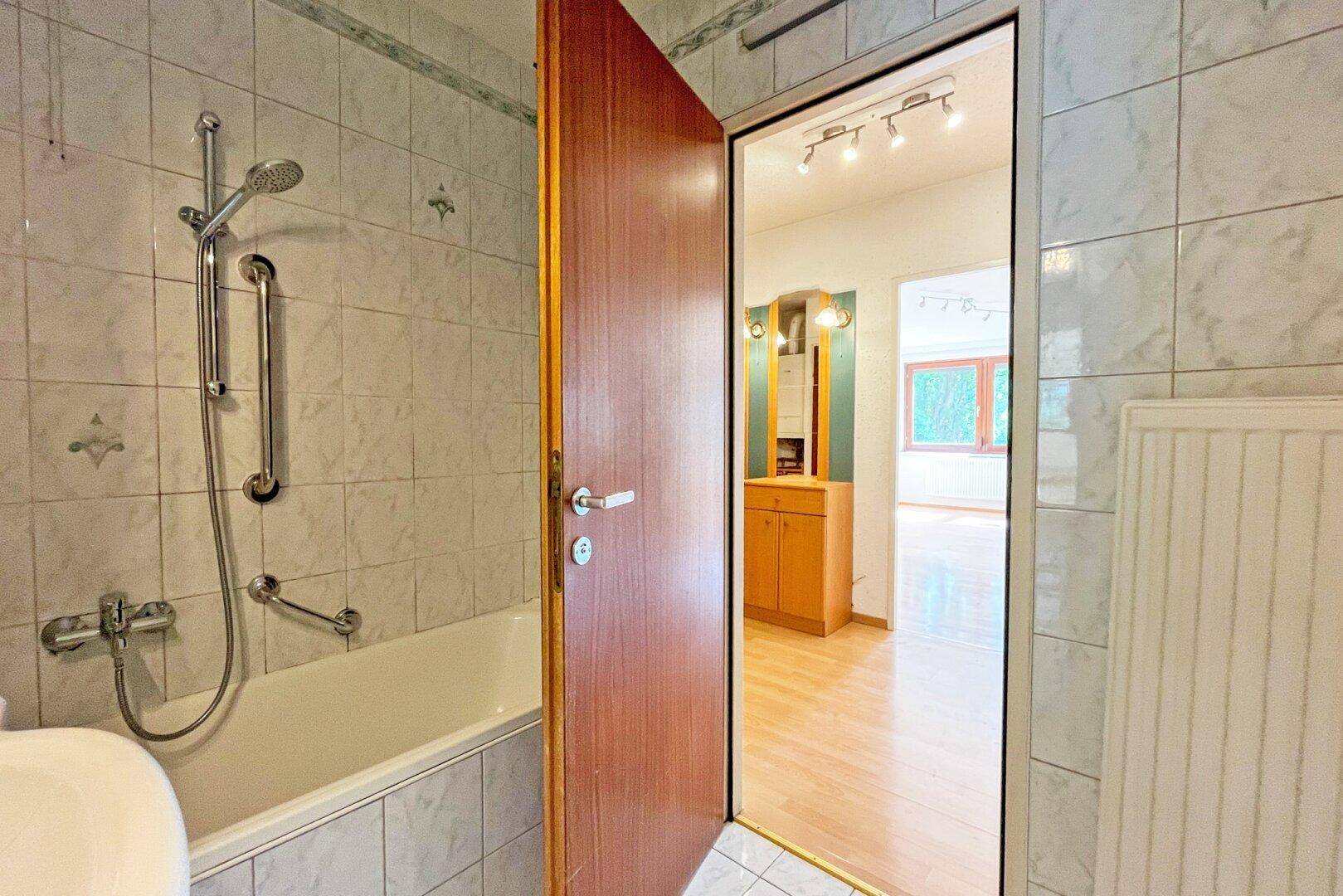 Badezimmer mit Badewanne, Duschfunktion und Haltegriffen, mit Blick in den angrenzenden Flur.