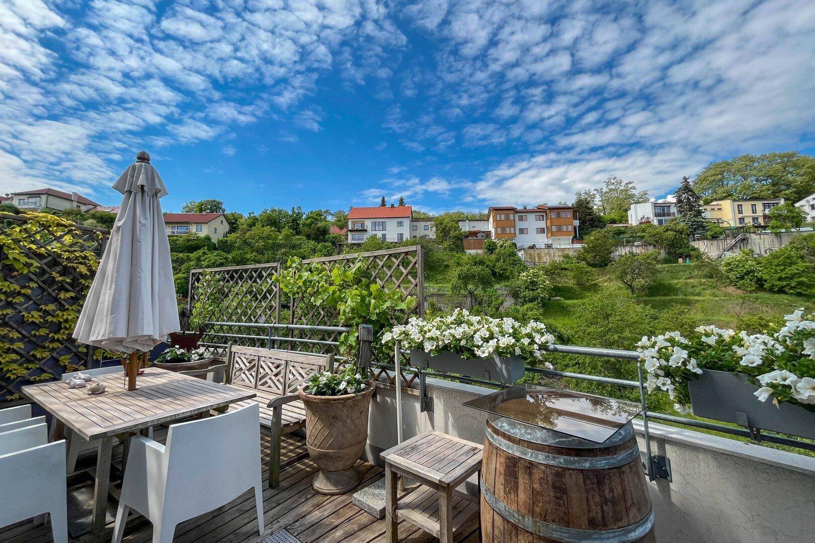 Möblierte Terrasse mit Holzboden, Sonnenschirm und Blick auf die grüne Umgebung unter blauem Himmel.