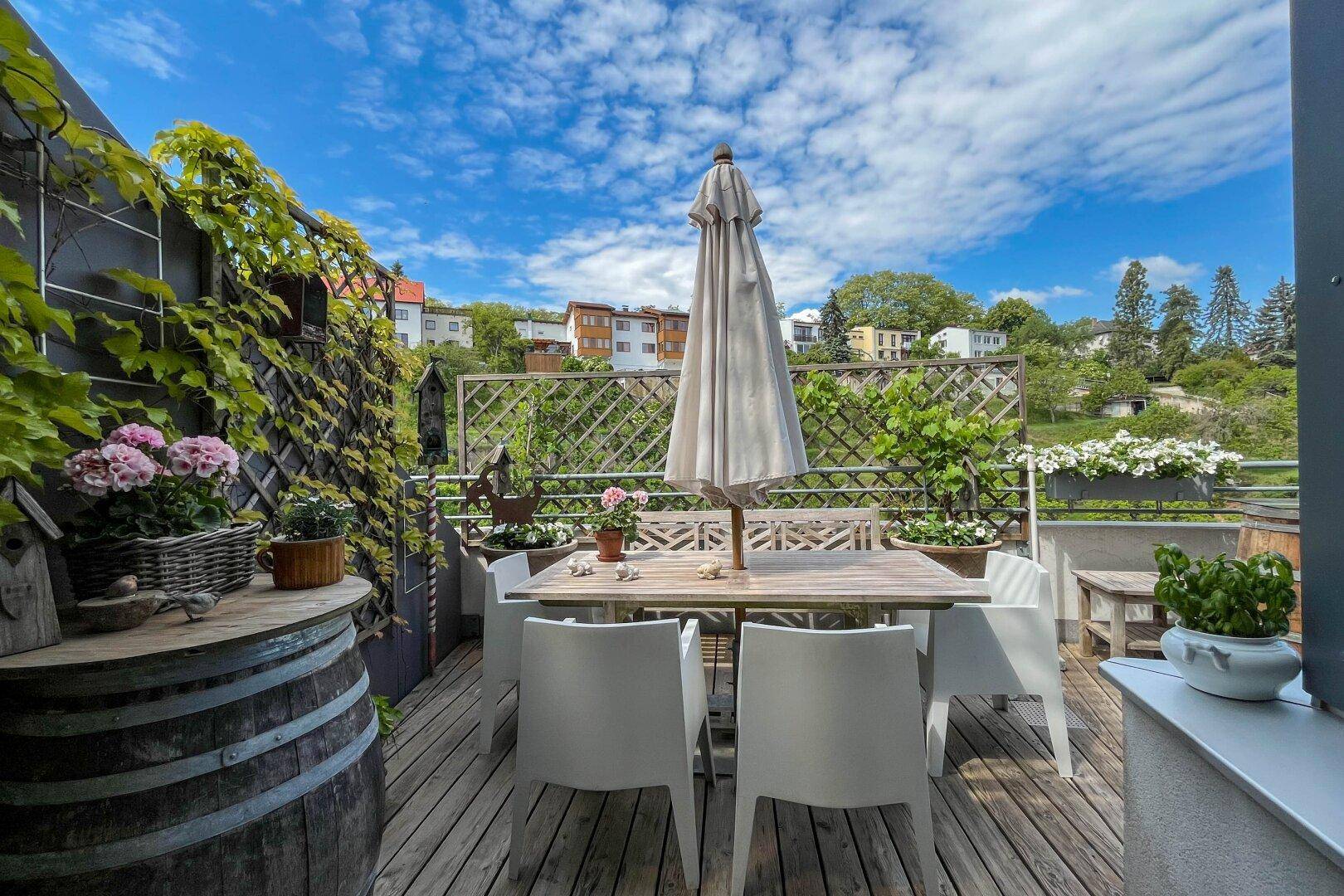 Gemütliche Terrasse mit Holztisch, Stühlen, Sonnenschirm und Pflanzen, umgeben von viel Grün.