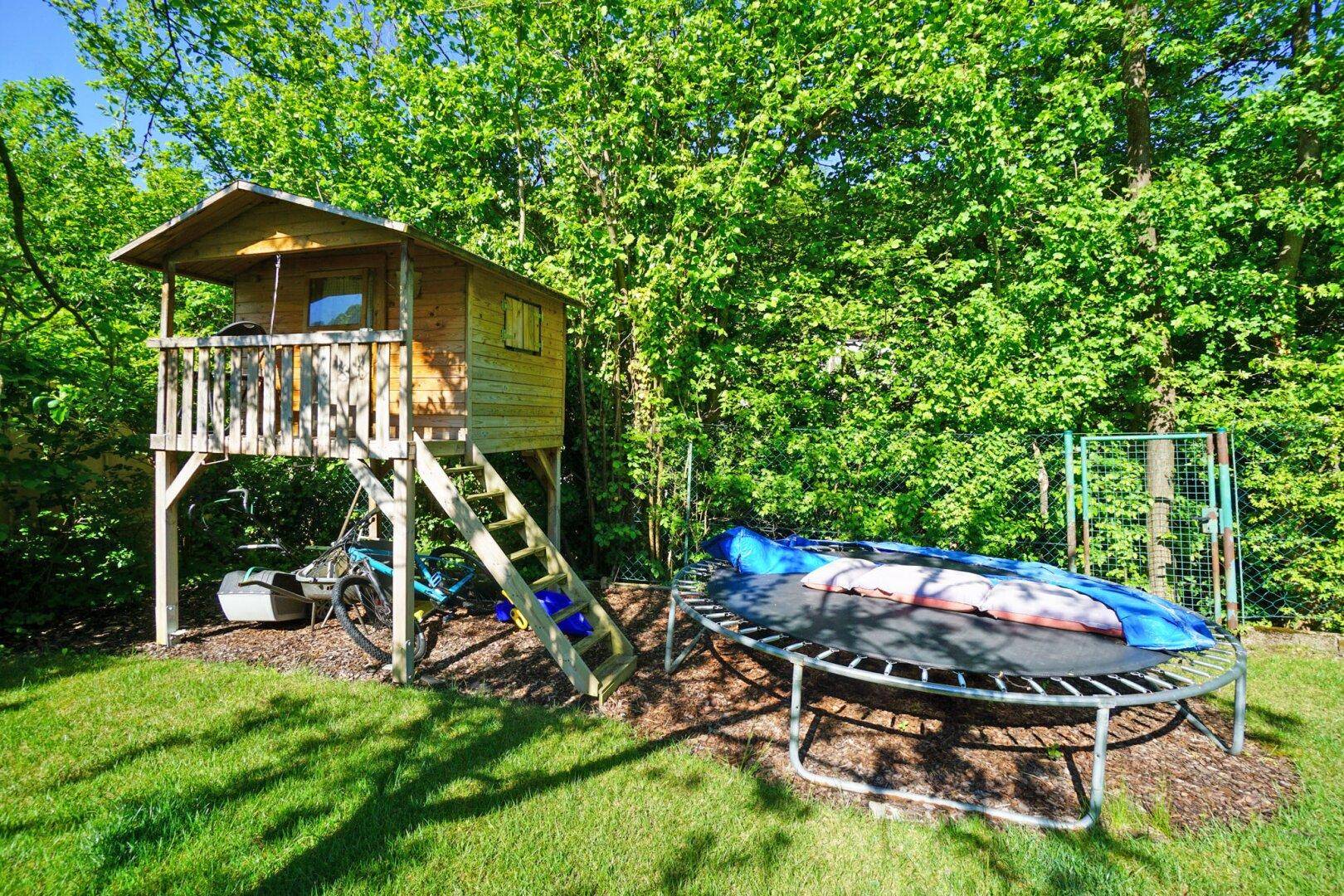 Spielbereich im Garten mit Baumhaus und Trampolin, umgeben von üppigem Grün.