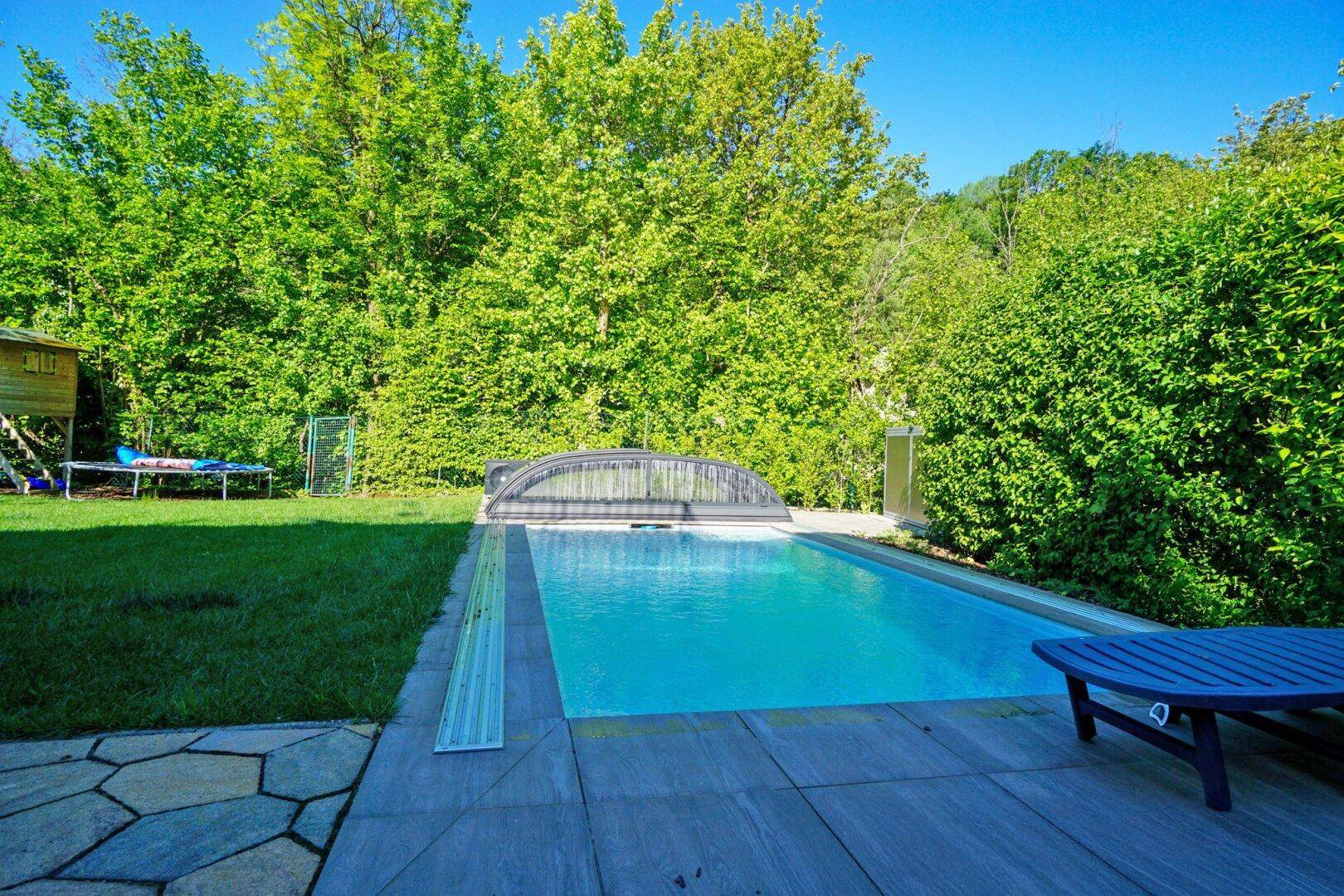 Poolbereich im Garten mit Liegestuhl und umgebender Hecke unter blauem Himmel.