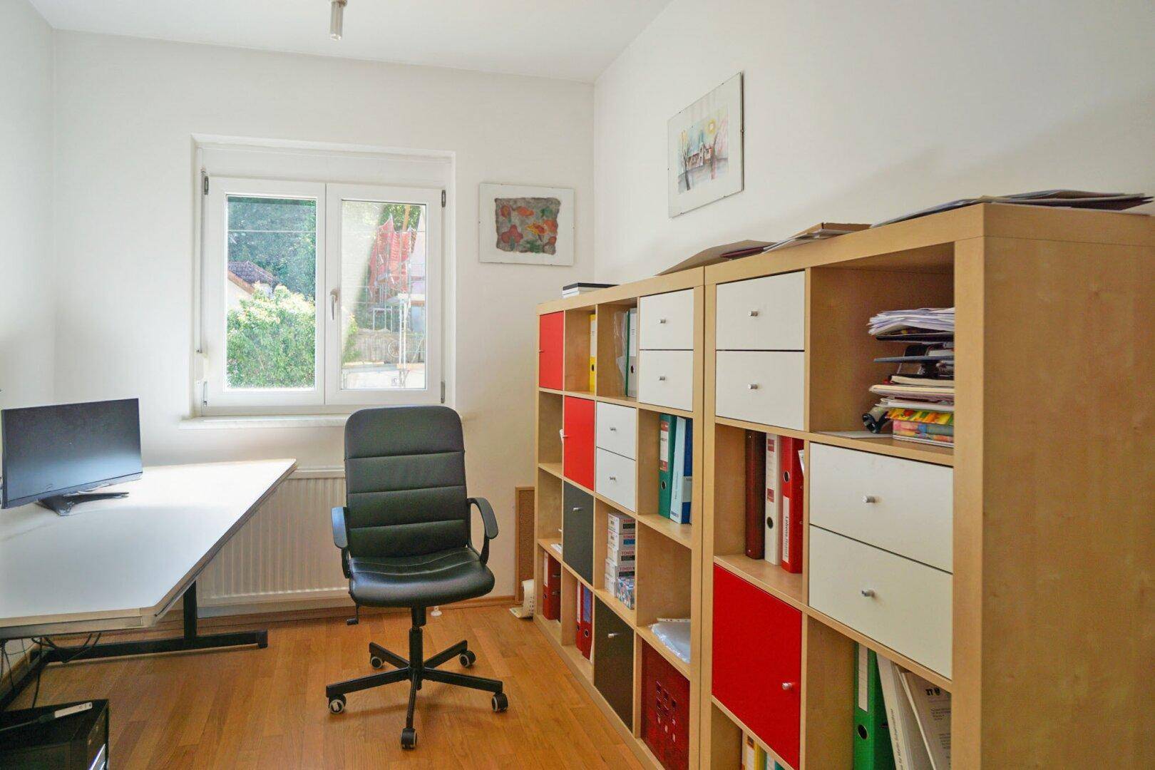Helles Arbeitszimmer mit Schreibtisch, Bürostuhl und Bücherregal mit Blick ins Grüne.