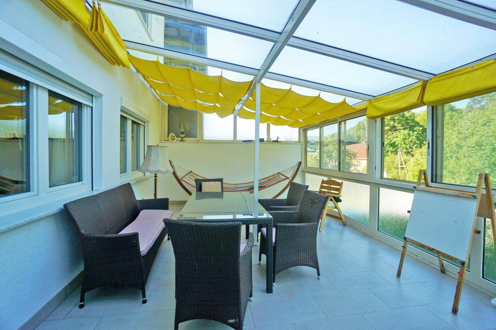 Heller Wintergarten mit Sitzmöbeln, Hängematte und Blick in die grüne Umgebung.