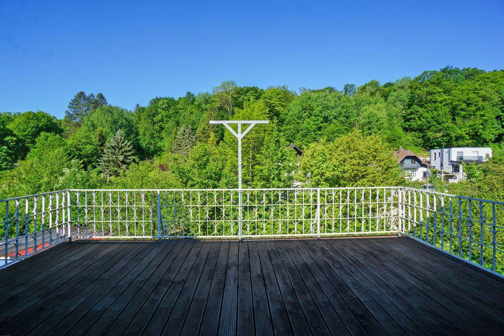 Großer Balkon mit Holzboden und schmiedeeisernem Geländer, umgeben von Bäumen.