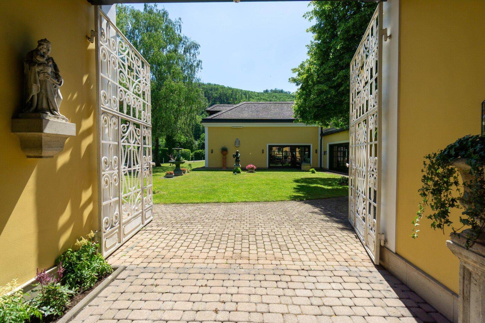Blick durch ein großes, weißes Tor auf den weitläufigen Garten mit Rasen und Bäumen.