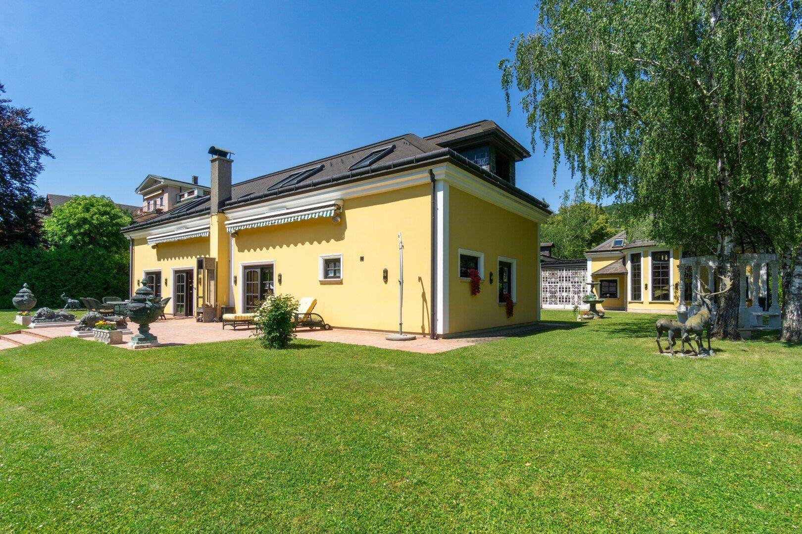 Seitenansicht des gelben Gebäudes mit Terrasse und weitläufigem Rasen, umgeben von viel Grün.