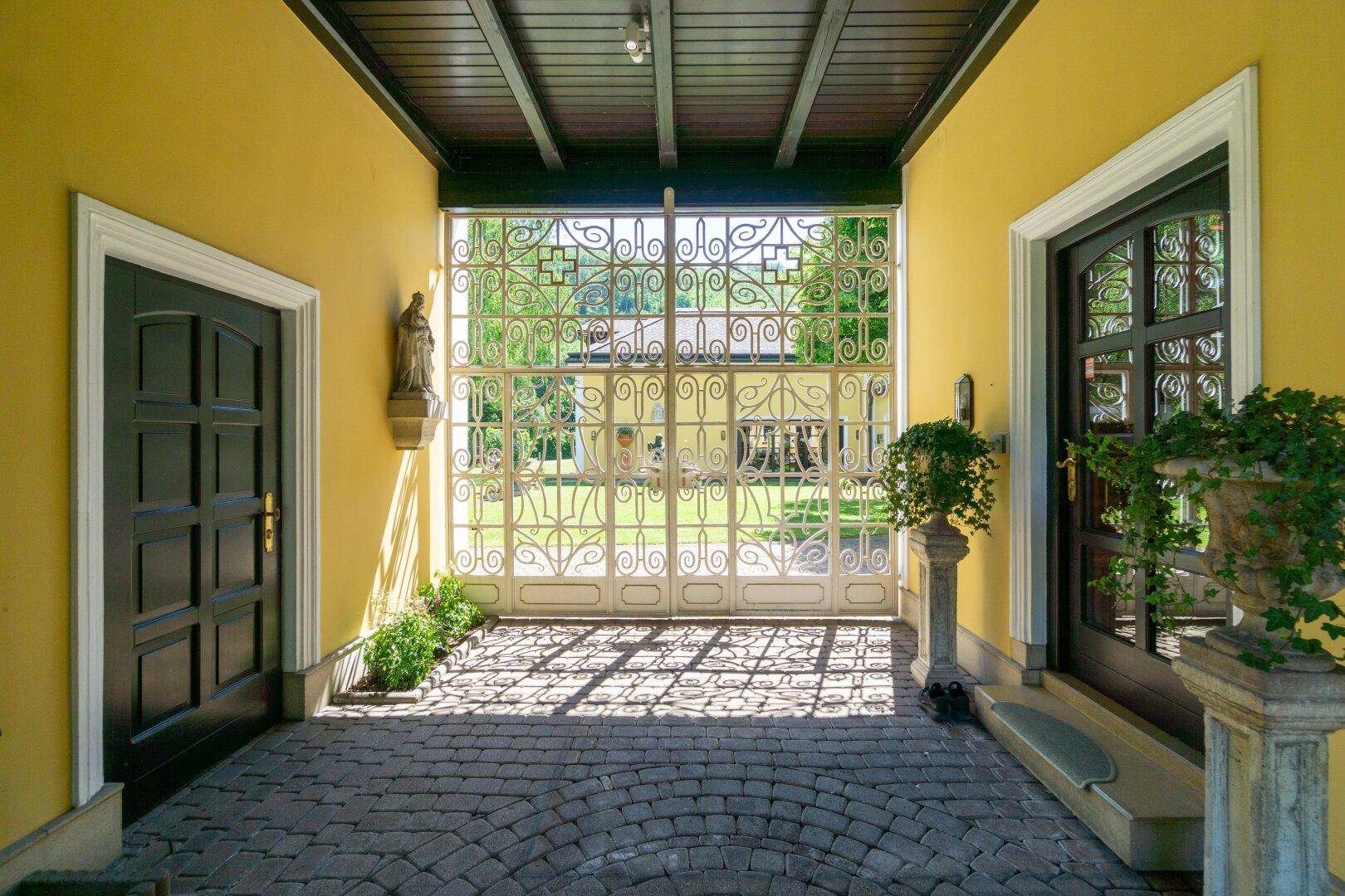 Überdachter Eingangsbereich mit gepflastertem Boden und schmiedeeisernem Tor, das zum Garten führt.
