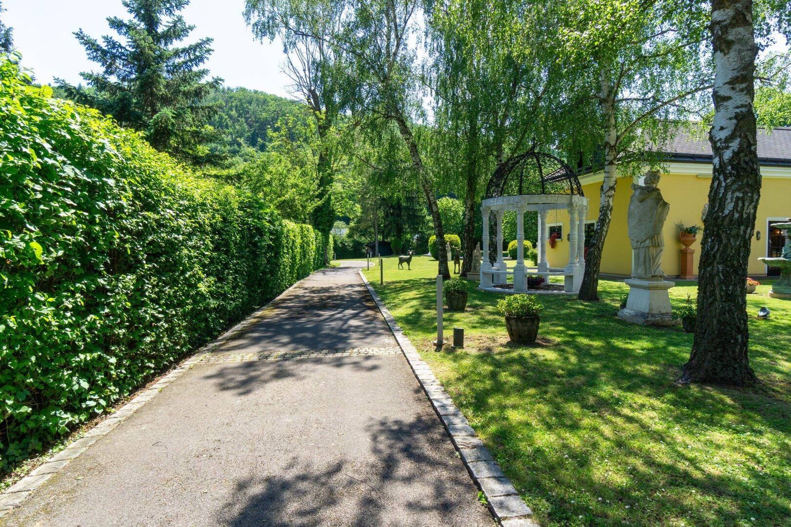 Gepflegter Gartenbereich mit Spazierweg, hohen Hecken, Bäumen und einem dekorativen Pavillon.