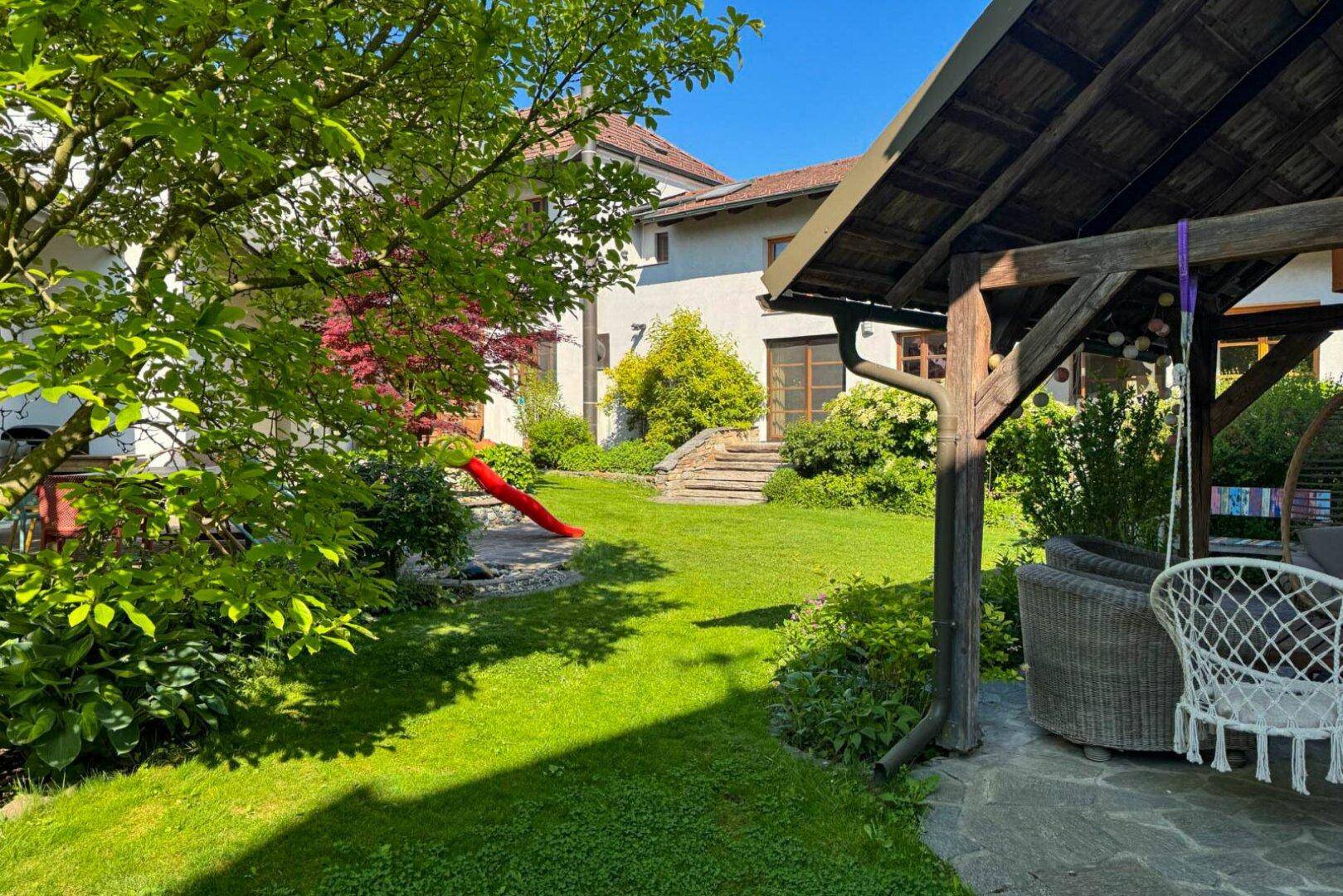 Sonniger Garten mit Spielrutsche, überdachtem Sitzbereich und viel Grünfläche.