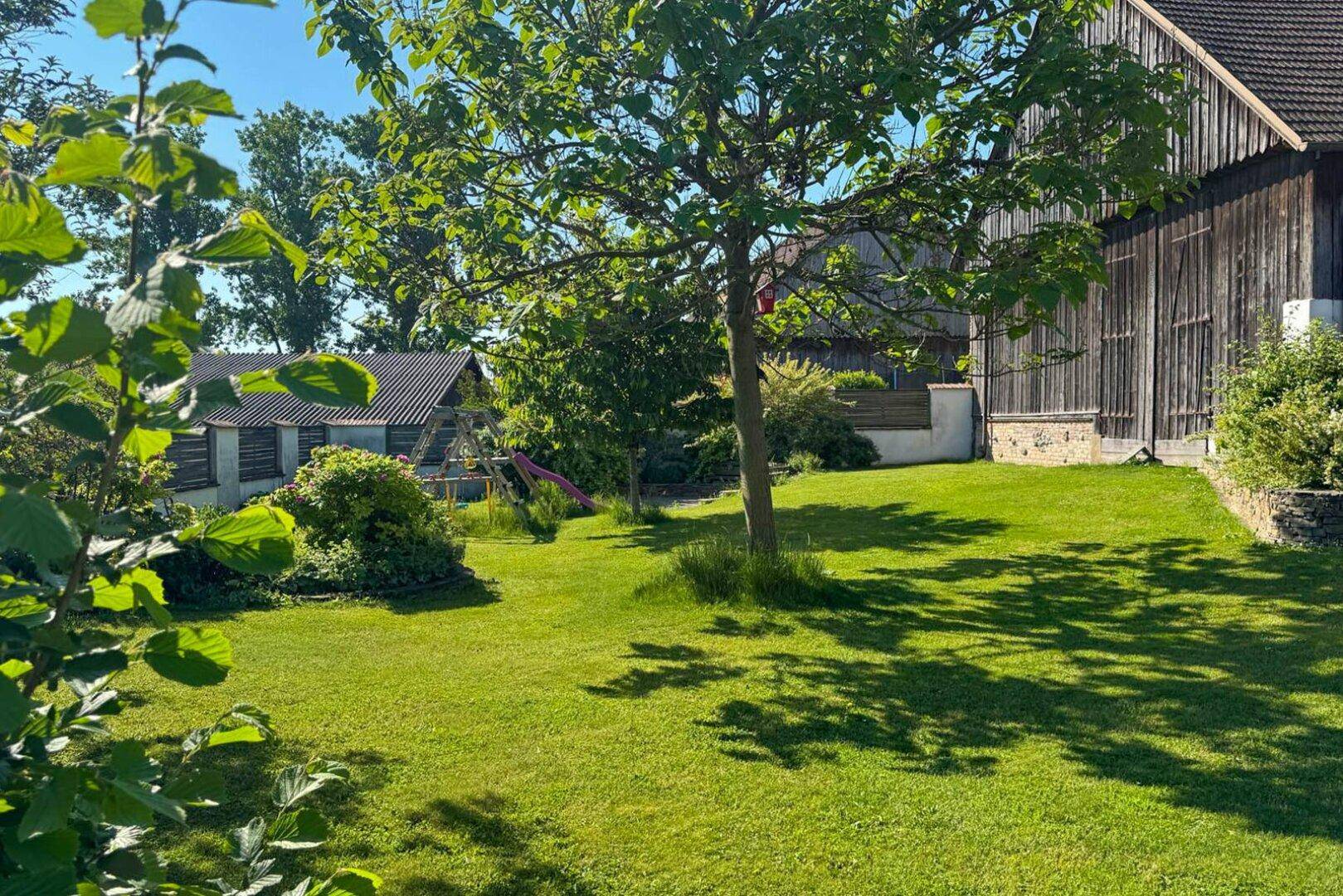 Weitläufiger Gartenbereich mit Rasen, Bäumen und Blick auf angrenzende Gebäude.