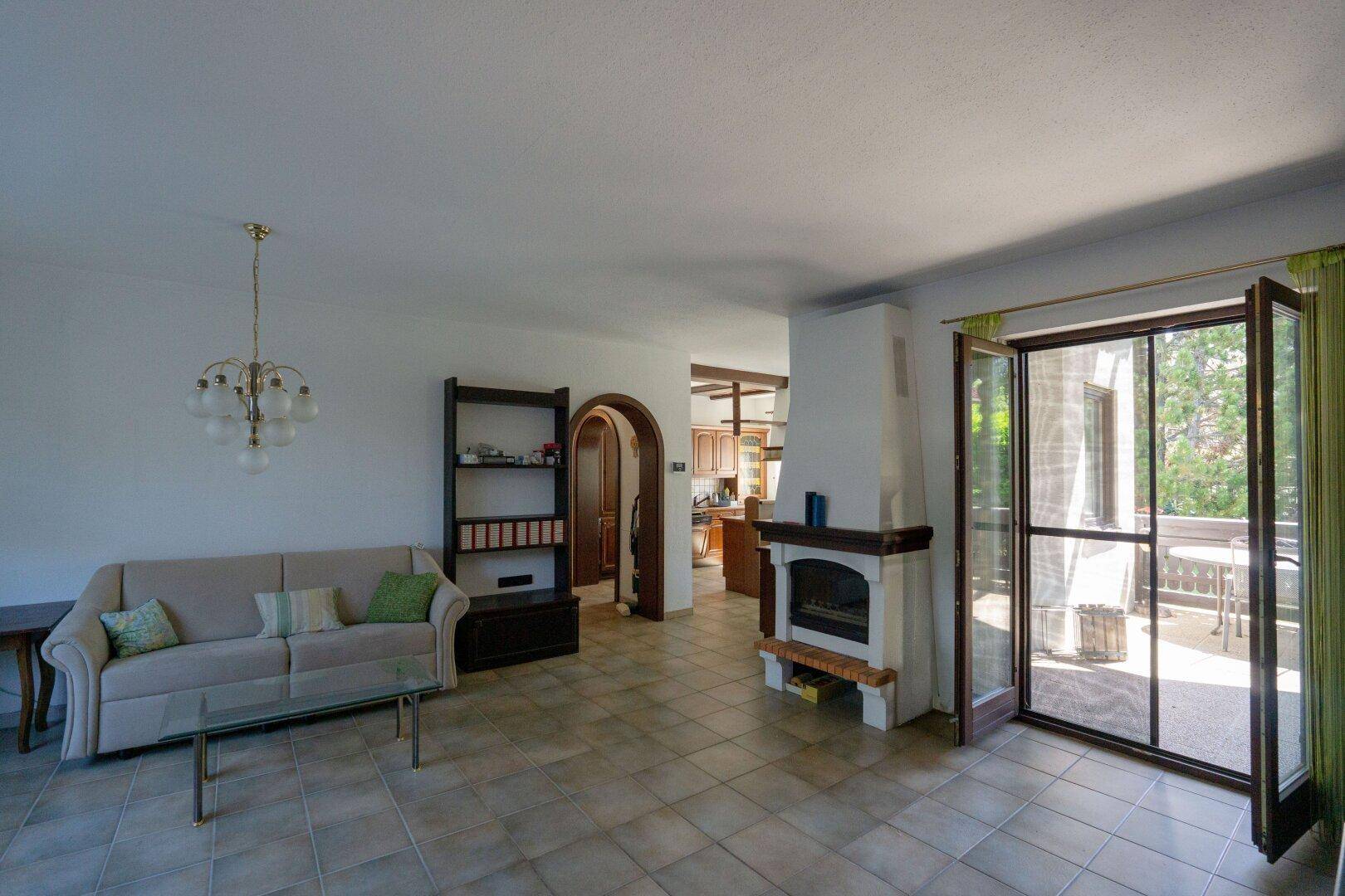 Helles Wohnzimmer mit beigem Sofa, Kamin und großen Fenstern zur Terrasse, gut beleuchtet.