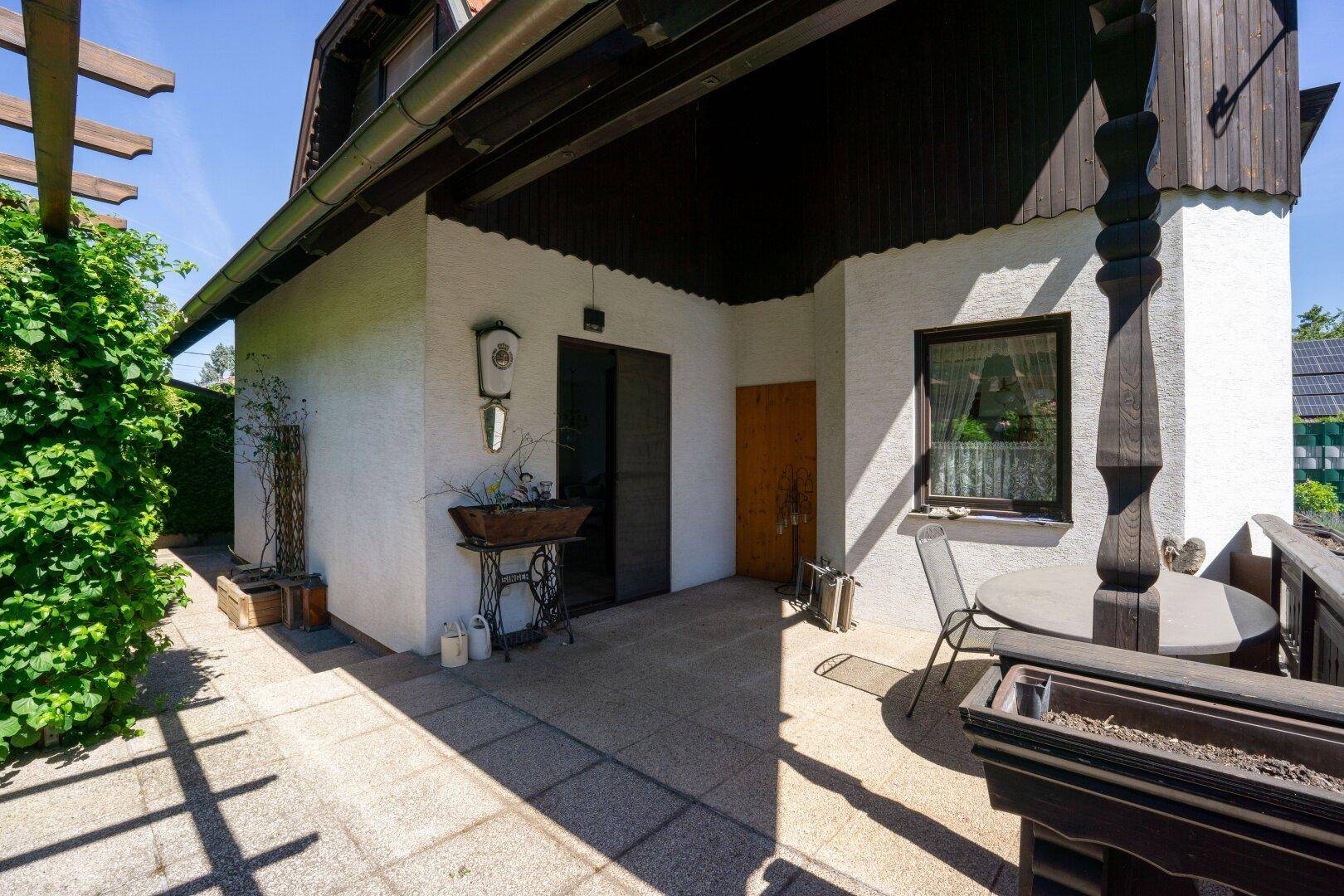 Überdachte Terrasse mit Steinfliesen und Zugang zum Haus, umgeben von Grünpflanzen und Ranken.