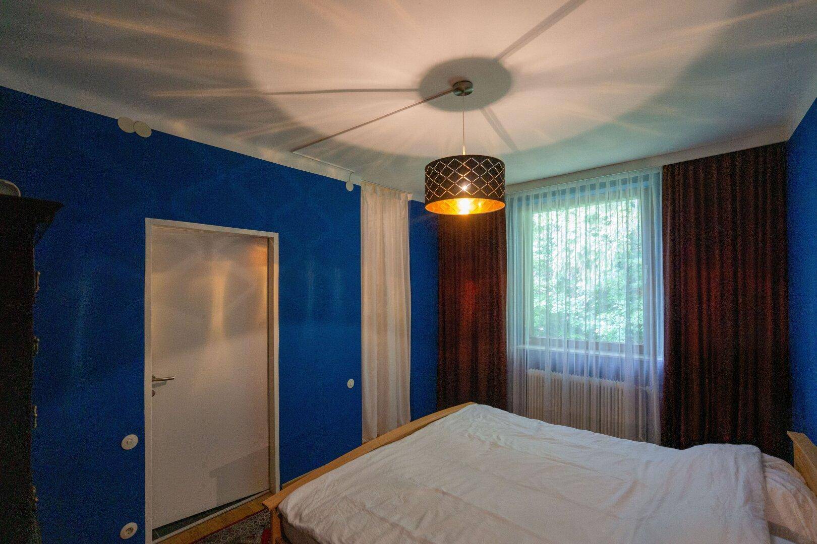 Schlafzimmer mit blauen Wänden, Doppelbett und Fenster mit Vorhängen.
