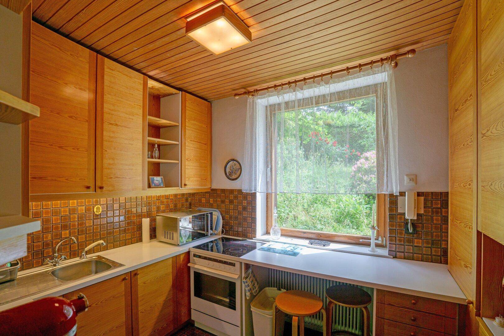 Kleine Küche mit Holzschränken, Fliesenrückwand und Fenster mit Blick ins Grüne.